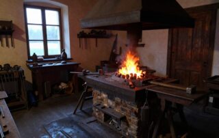Weitwinkelaufnahme einer deutschen Schmiedewerkstatt im Morgengrauen: alte Schmiede, glühende Kohlen, Hammer auf der Werkbank und Fenster mit ersten Lichtstrahlen
