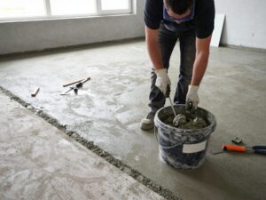 Bodenleger bereitet Betonuntergrund für Ausgleich vor, trägt Schutzbrille und Handschuhe, steht neben Mischbehälter mit Ausgleichsmasse