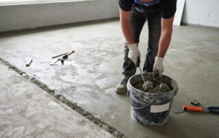 Bodenleger bereitet Betonuntergrund für Ausgleich vor, trägt Schutzbrille und Handschuhe, steht neben Mischbehälter mit Ausgleichsmasse