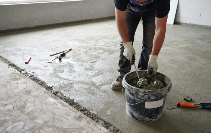 Bodenleger bereitet Betonuntergrund für Ausgleich vor, trägt Schutzbrille und Handschuhe, steht neben Mischbehälter mit Ausgleichsmasse