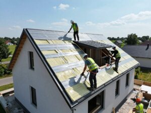 Arbeiter in Schutzkleidung installieren Hochleistungsdämmplatten auf einem schrägen deutschen Wohnhausdach unter klarem Himmel