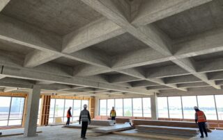 Modernes Baustellenbild mit teilweise fertiggestellter Decke aus Betonbalken, Holzbalken und Gipskartonplatten, Arbeiter tragen Helme und Warnwesten