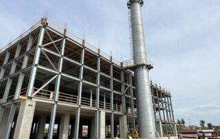 Weitwinkelaufnahme einer modernen deutschen Industriehalle im Bau, Stahlrahmen-Schornstein und Betonfundamente, Arbeiter in Sicherheitswesten prüfen die Struktur