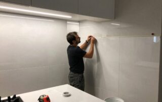 Mitarbeiter legt Malerband an Kachelwand, um Fuge zu versiegeln – LED-Beleuchtung, Messwerkzeug und Kleister im Vordergrund