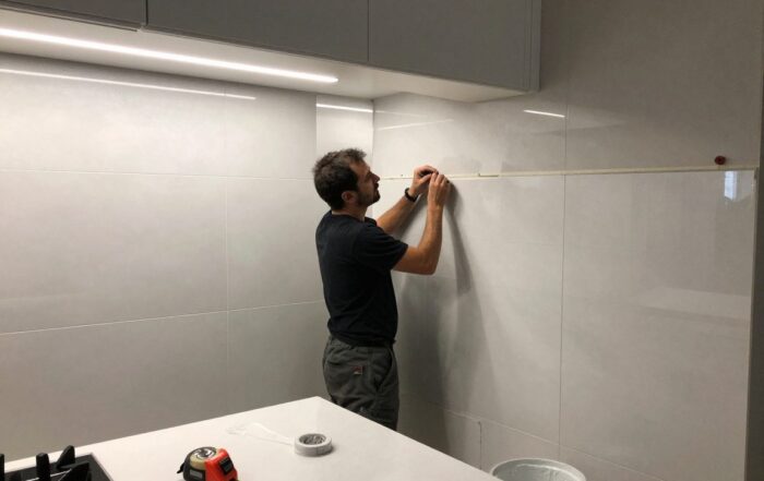 Mitarbeiter legt Malerband an Kachelwand, um Fuge zu versiegeln – LED-Beleuchtung, Messwerkzeug und Kleister im Vordergrund