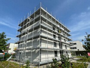 Weitwinkelaufnahme eines modernen Metallgerüsts an der Fassade eines deutschen Einfamilienhauses, mit klaren blauen Himmel und umgebender Grünfläche, mehreren Ebenen, Sicherheitsgeländern und Arbeitern in Helmen