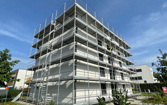 Weitwinkelaufnahme eines modernen Metallgerüsts an der Fassade eines deutschen Einfamilienhauses, mit klaren blauen Himmel und umgebender Grünfläche, mehreren Ebenen, Sicherheitsgeländern und Arbeitern in Helmen