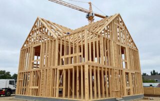 Weitwinkelaufnahme eines modernen Holzrahmenhauses in Bauphase mit freiliegenden Balken und Pfosten, Kranen und Arbeitern im Vordergrund