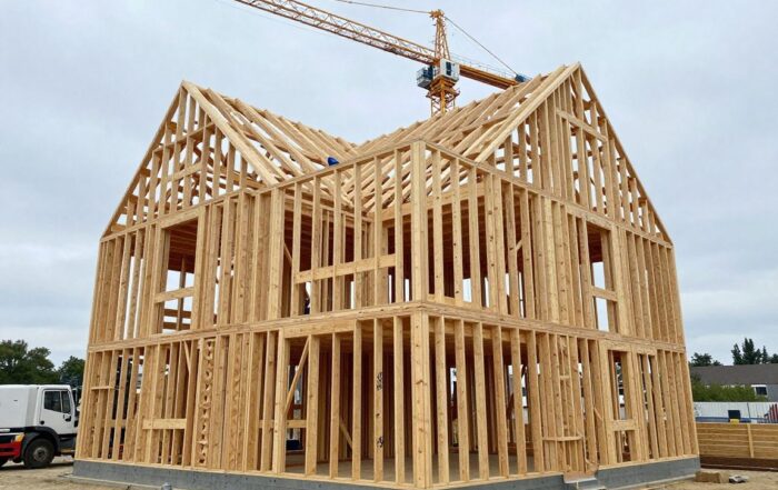 Weitwinkelaufnahme eines modernen Holzrahmenhauses in Bauphase mit freiliegenden Balken und Pfosten, Kranen und Arbeitern im Vordergrund