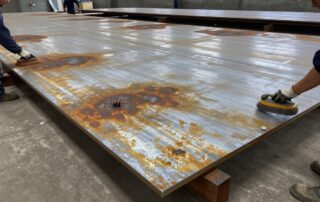 Große Stahlplatte mit Rostflecken und scharfen Kanten, die von Arbeitern in Schutzhelm und Handschuhen mit Bürsten und Schleifwerkzeugen vorbereitet wird