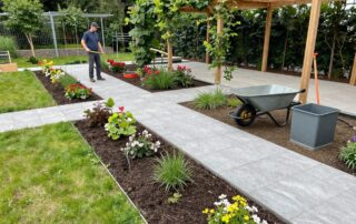 Breiter Blick auf einen deutschen Garten im Bau: Natursteinwege, frisch gepflanzte Blumenbeete und eine Holzwand mit Kletterpflanzen. Ein Landschaftsarchitekt hält ein Maßband, umgeben von Werkzeugen wie Spaten, Schubkarren und einem kleinen Kompostbehälter.