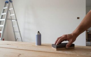 Hand hält Schleifblock auf Holzwand, neben Sprühdose und Malerleiter an Baustelle