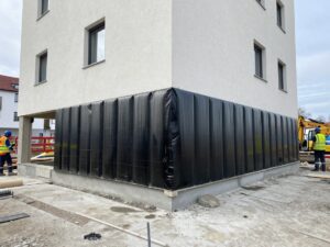 Kellerwand mit vertikaler Feuchtigkeitssperre aus Bitumenmembran, umgeben von Baugeräten und Arbeitern in Schutzkleidung