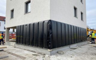 Kellerwand mit vertikaler Feuchtigkeitssperre aus Bitumenmembran, umgeben von Baugeräten und Arbeitern in Schutzkleidung