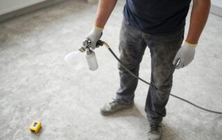 Professioneller Bodenleger steht auf Beton, hält Sprühgerät und Grundierungsflasche, trägt Handschuhe und Schutzbrille