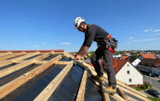 Deutscher Dachdecker im Sicherheitshelm und -gurt, der Edelstahlschrauben an einer Schrägdachfläche mit Bitumenmembran befestigt