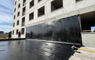 Arbeiter legen Bitumenbahne auf die Außenseite einer modernen deutschen Wohnfassade, Stahlbewehrung sichtbar durch das Material