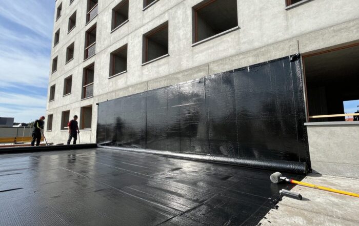 Arbeiter legen Bitumenbahne auf die Außenseite einer modernen deutschen Wohnfassade, Stahlbewehrung sichtbar durch das Material