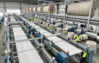 Breitwinkelaufnahme eines industriellen Seilwerkes mit Maschinen, die synthetische Fasern zu Seilen verarbeiten, während Arbeiter Schutzwesten tragen und Bündel inspizieren