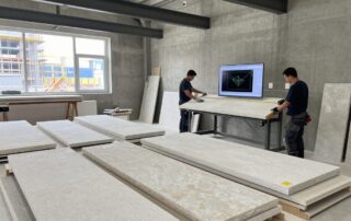 Weitwinkelaufnahme eines deutschen Steinmetzbetriebs mit CAD-Software auf einem Tisch, Steinplatten und Handwerkern in Arbeitsglehn