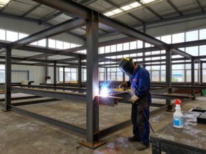 Schweißer in Schutzkleidung entfernt Staub und Rost von einer Schweißnaht mit Sprühreiniger, während ein Stahlrahmen unter Konstruktion steht