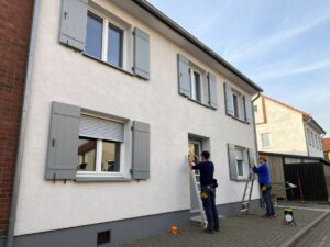 Handwerker messen an einer deutschen Hausfassade mit Maßband und Lasermessgerät die Position von Rolläden, während eine Leiter im Bild zu sehen ist.