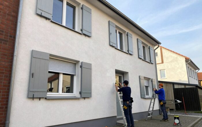 Handwerker messen an einer deutschen Hausfassade mit Maßband und Lasermessgerät die Position von Rolläden, während eine Leiter im Bild zu sehen ist.