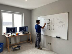 Techniker zeichnet mit Bleistift einen Sanitärplan auf einem großen Whiteboard, umgeben von Werkzeugen und CAD-Monitoren im hellen Arbeitsraum