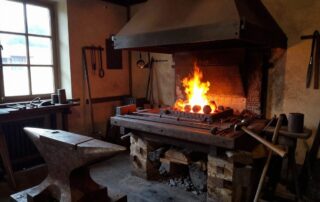 Weitwinkelaufnahme einer traditionellen deutschen Schmiede mit glühenden Kohlen, Amboss, Werkbank und Werkzeugen, warmes Licht aus der Schmiedefeuerung