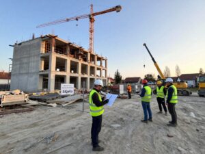 Sicherheitskoordinator steht bei einem frühen Morgen an einer deutschen Abrissstelle, umgeben von Sicherheitszaun, Beschilderung und Baustellenpersonal mit Helmen und Reflektoren.