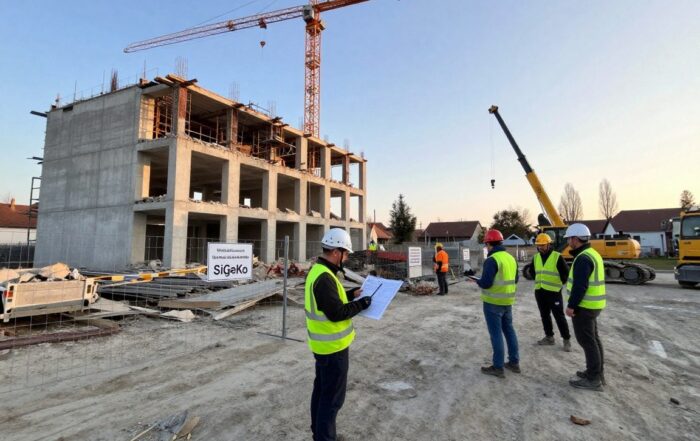 Sicherheitskoordinator steht bei einem frühen Morgen an einer deutschen Abrissstelle, umgeben von Sicherheitszaun, Beschilderung und Baustellenpersonal mit Helmen und Reflektoren.