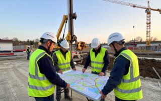 Team von Spezialbohrern in Sicherheitshelmen und Warnwesten steht um eine detaillierte topografische Karte auf einem Klapptisch, während im Hintergrund ein moderner Bohrturm neben einer teilweise ausgegrabenen Grube steht.