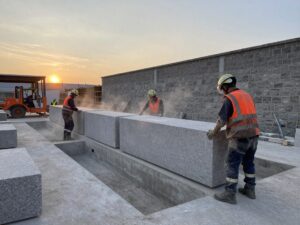 Großaufnahme einer deutschen Steinmetzwerkstatt im Abendlicht: Arbeiter in Schutzkleidung bearbeiten Granitblöcke, moderne Maschinen und traditionelle Mauerarbeiten sind sichtbar.
