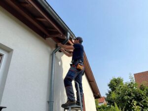 Erfahrener Spengler legt ein Metall-Fallrohr an die Dachrinne eines traditionellen deutschen Hauses, umgeben von Grün und blauem Himmel.