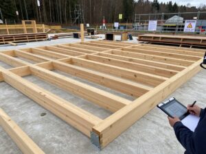 Weitwinkelaufnahme eines deutschen Holzbauprojekts mit teilweise montierten Balken auf Betonfundament, Gerüst und Sicherheitsbeschilderung