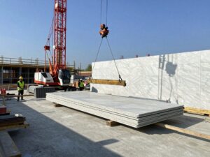 Hydraulische Kräne heben große Marmorplatten auf eine Betonfundament, Arbeiter in Helmen und reflektierenden Westen arbeiten unter klarem blauen Himmel.