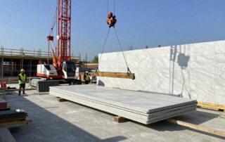 Hydraulische Kräne heben große Marmorplatten auf eine Betonfundament, Arbeiter in Helmen und reflektierenden Westen arbeiten unter klarem blauen Himmel.