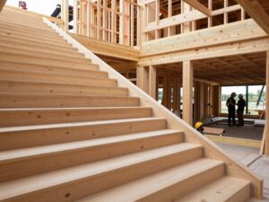 Weitwinkelaufnahme einer modernen Holztreppe auf einem deutschen Baustellenplatz mit Holzbalken, Stahlbrackets und Arbeitern in Helmen unter natürlichem Licht